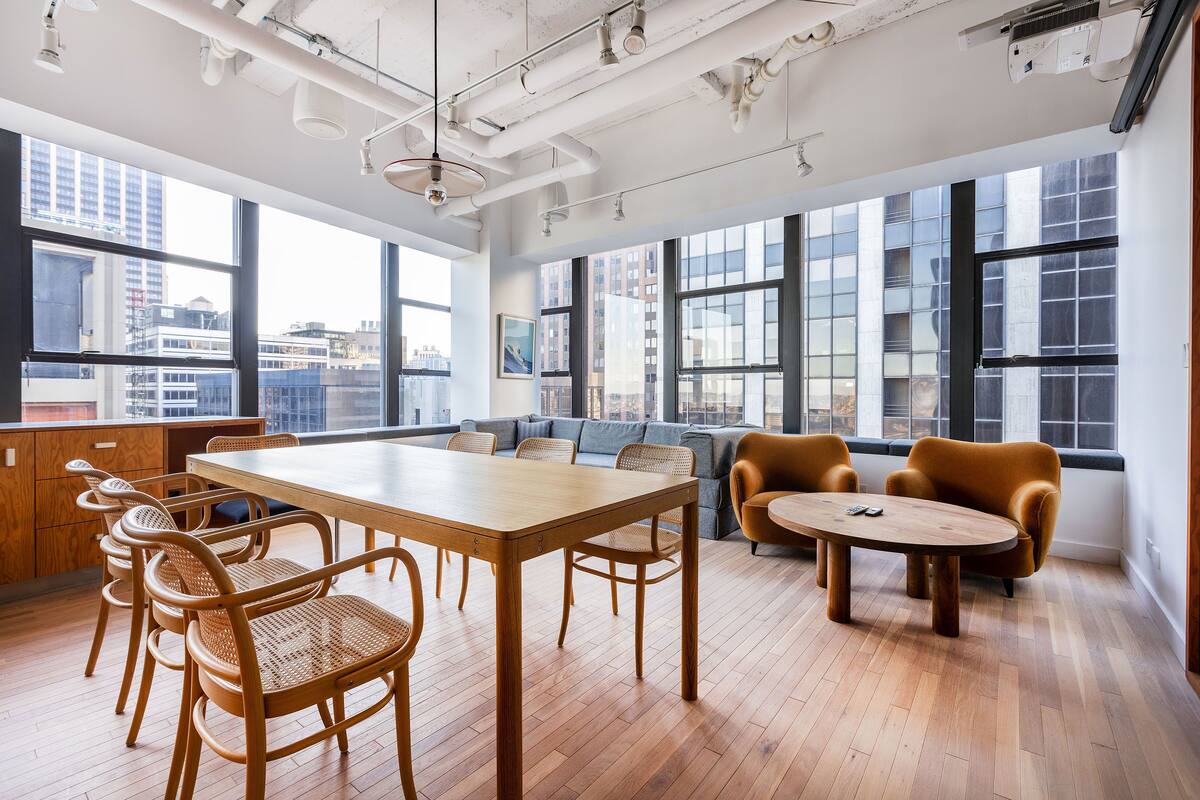 A spacious office with a wooden table and chairs, a sofa, and large windows offering city views.