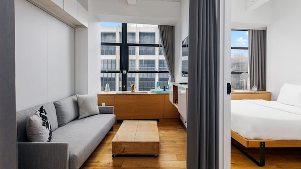 Una habitación de hotel moderna con sofá, una mesita de centro de madera, cama y escritorio con silla. Amplias ventanas ofrecen vistas de la ciudad.