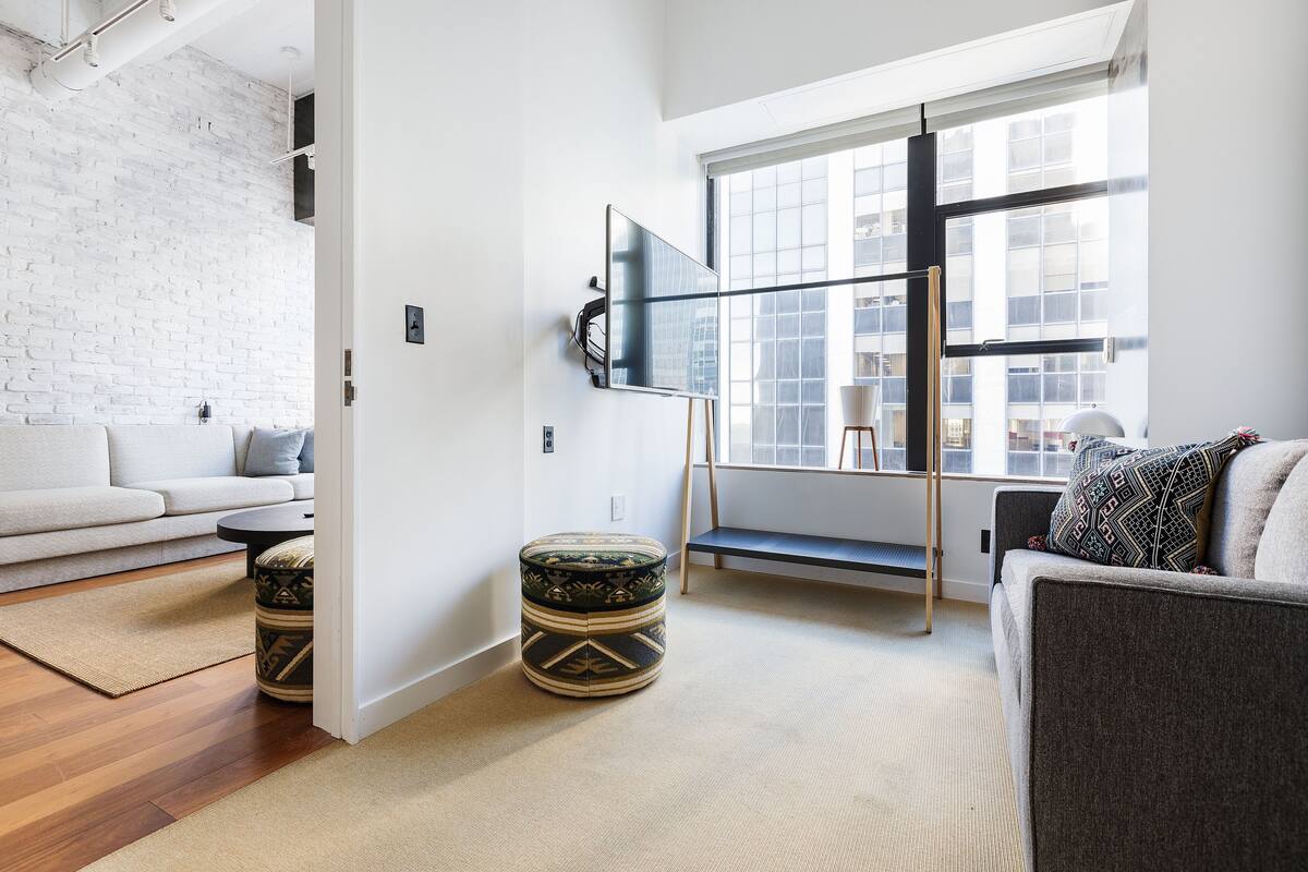 A modern living room with a white brick wall, a large window, a grey sofa, a round ottoman, and a mounted TV.