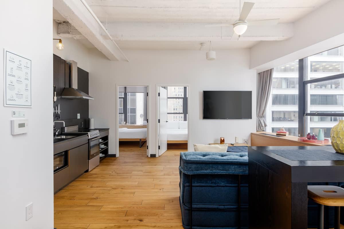 A modern kitchen with wooden flooring, a dining area with a blue cushioned bench, a TV mounted on the wall, and a large window with a city view.