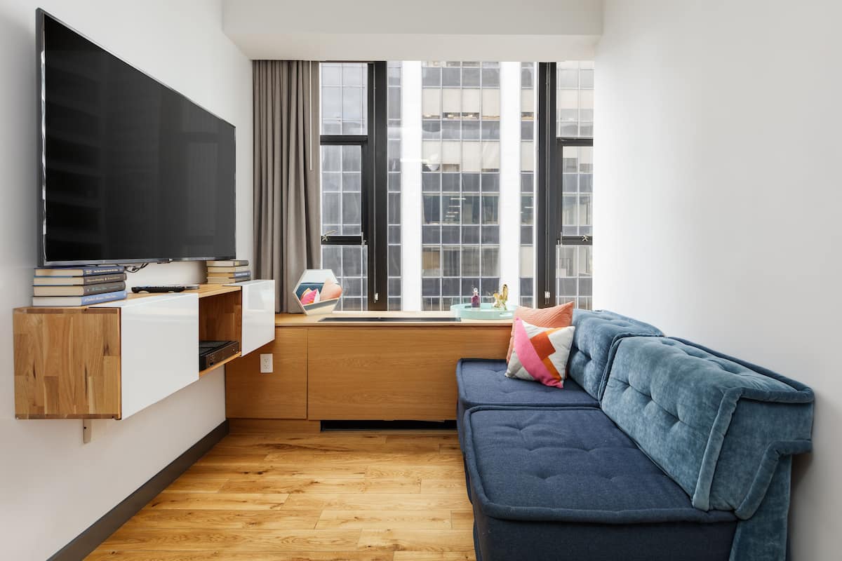 A modern living room with a large flat-screen TV mounted on the wall, a wooden cabinet below it, a blue sofa with cushions, and a window with a view of buildings.