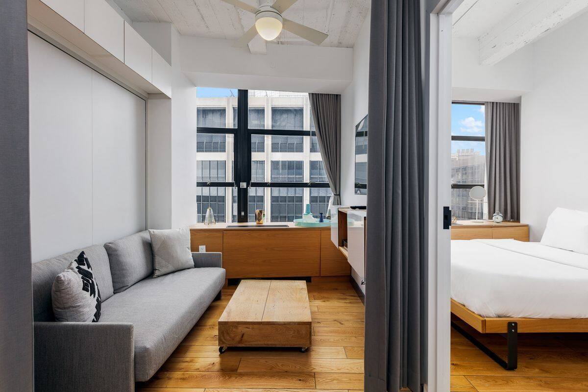 A modern hotel room with a sofa, a bed, a wooden coffee table, and a view of the city through large windows.