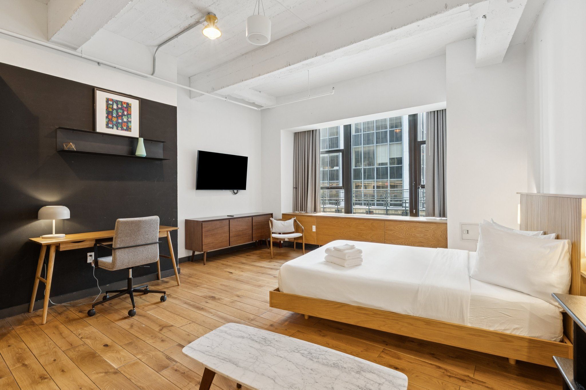 Ein modernes Hotelzimmer mit einem hölzernen Bett, einem Schreibtisch mit Stuhl, einem an der Wand befestigten Fernseher und einem großen Fenster mit Blick auf die Stadt.