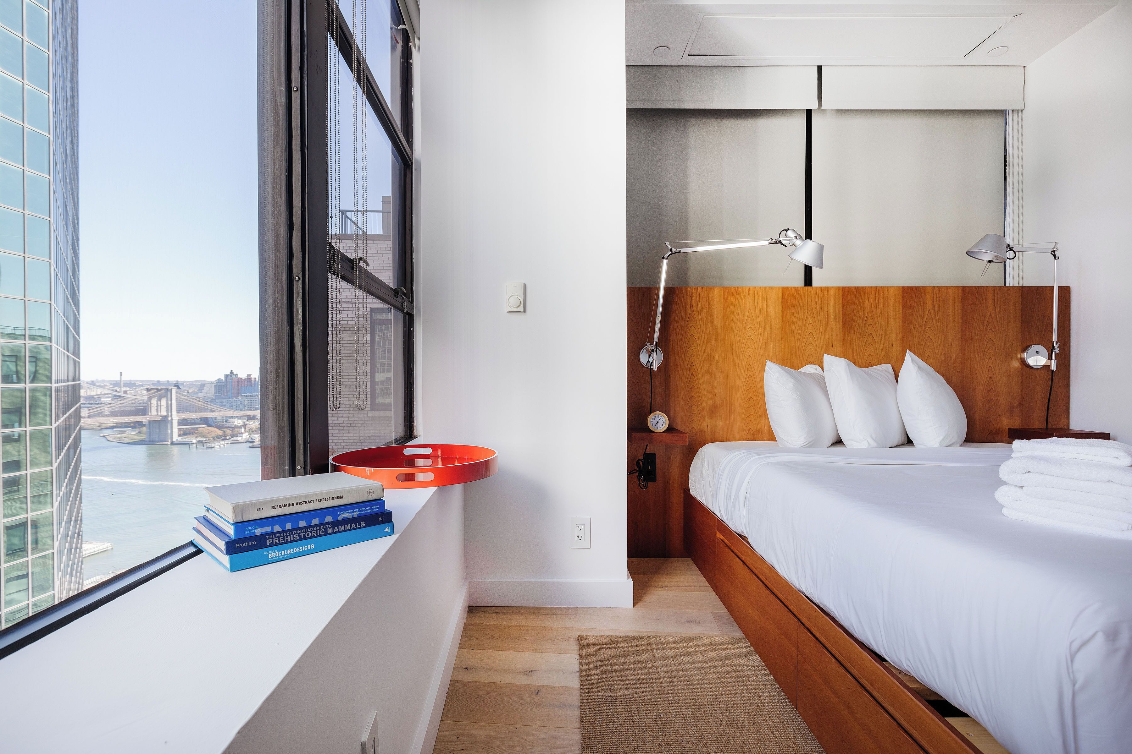 Een moderne hotelkamer met een groot raam, een houten hoofdbord, een nachtkastje met boeken en een rode dienblad.