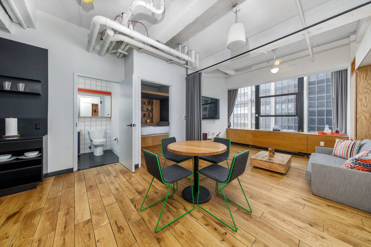A modern living space with a wooden floor, a round dining table with chairs, a grey sofa, and a TV mounted on the wall.