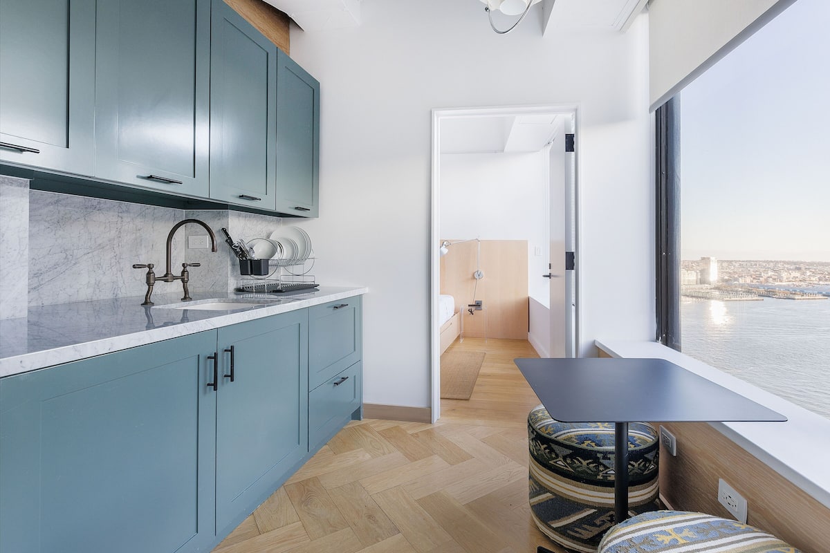 A modern kitchen with teal cabinets, a marble countertop, and a view of the city through a large window.