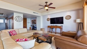 A spacious living room with a sofa, coffee table, and TV. There is a ceiling fan, a dining area in the background, and a kitchen visible through an open door.