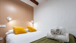 A hotel room with a wooden panel wall, a bed with white bedding and two yellow pillows, a green throw blanket, and a stack of white towels. - Apartment in TGN historic center only 450m beach (Tarragona)