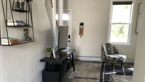Um quarto espaçoso com piso de madeira escura, uma escrivaninha preta com um computador, uma cadeira e uma janela com vista para o verde.