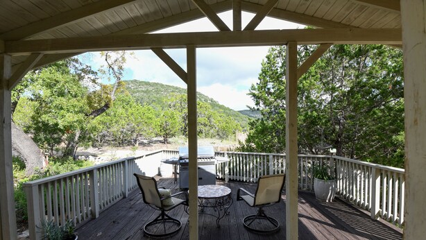 A covered deck with two chairs and a table, surrounded by a white railing, overlooking a natural landscape with trees and hills.