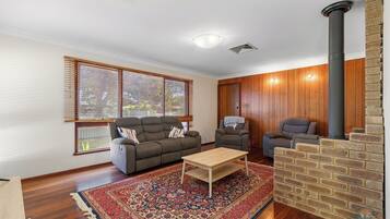 A living room with a sofa, two armchairs, a coffee table, a patterned rug, a brick fireplace, and wooden walls.
