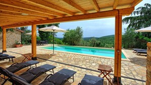 A covered outdoor swimming pool area with lounge chairs and umbrellas, surrounded by stone flooring and wooden beams. - Villa Collina - Holiday House Rental with private swimming pool in the small  village of Montegi... (Montegiove)