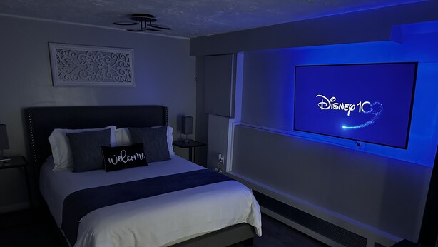Habitación de hotel moderna con una cama grande, un televisor montado en la pared mostrando el logo de Disney 1C, un ventilador de techo y un panel decorativo en la pared.