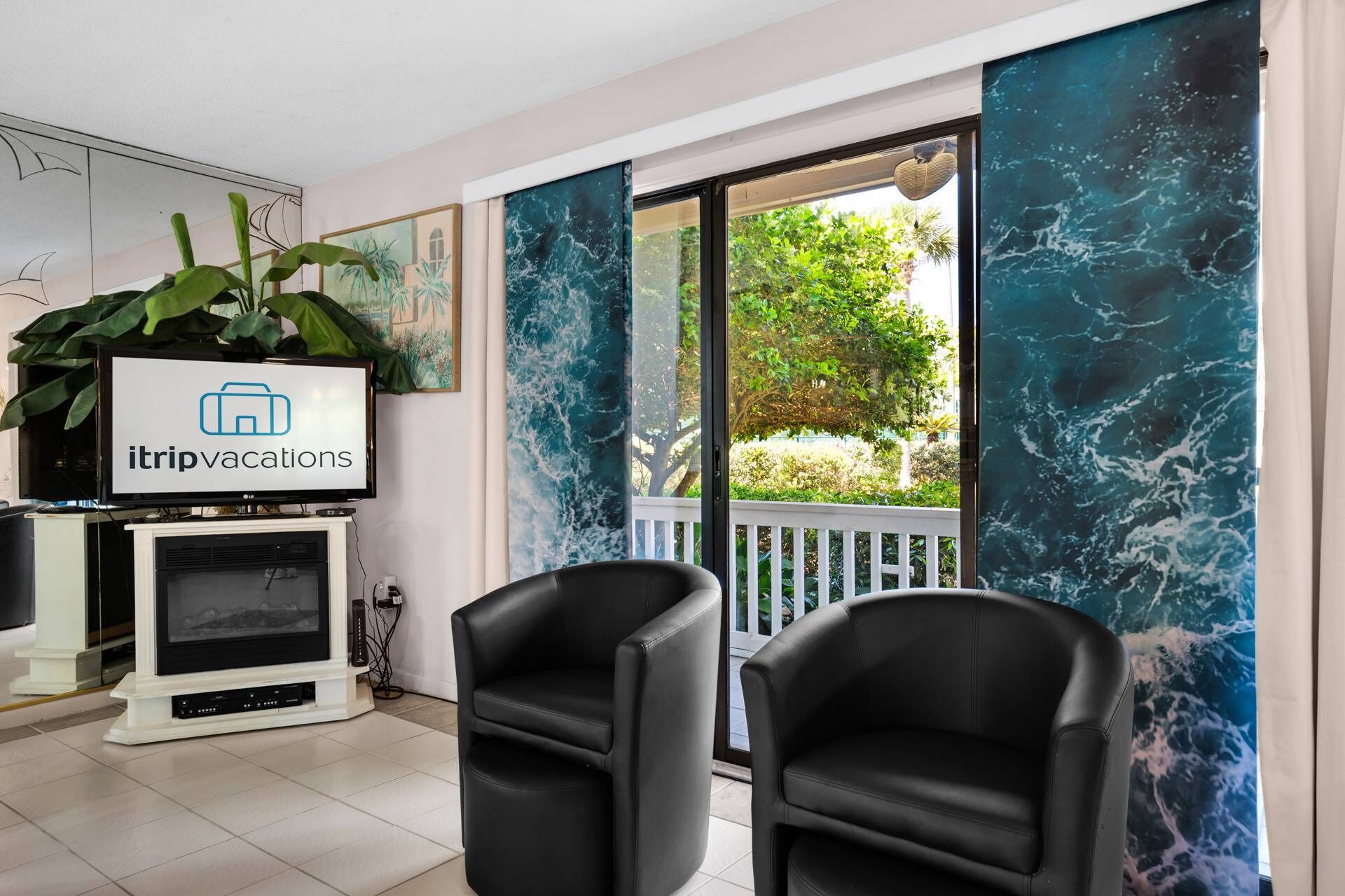 A living room with a television displaying 'itrip vacations', a large window with a view of greenery, and two black chairs.