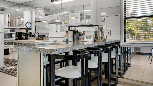 A modern kitchen with a granite countertop, stainless steel appliances, and a bar area with black stools. - Ground Floor Condo | 2-Min Walk to Beach | Pools | Tennis | Shuffleboard | Sleeps 4 | WiFi! (Santa Rosa Beach)