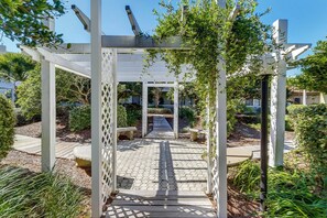 Ein überdachter Gartenweg mit einer Pergola, umgeben von Grünpflanzen und blauem Himmel.