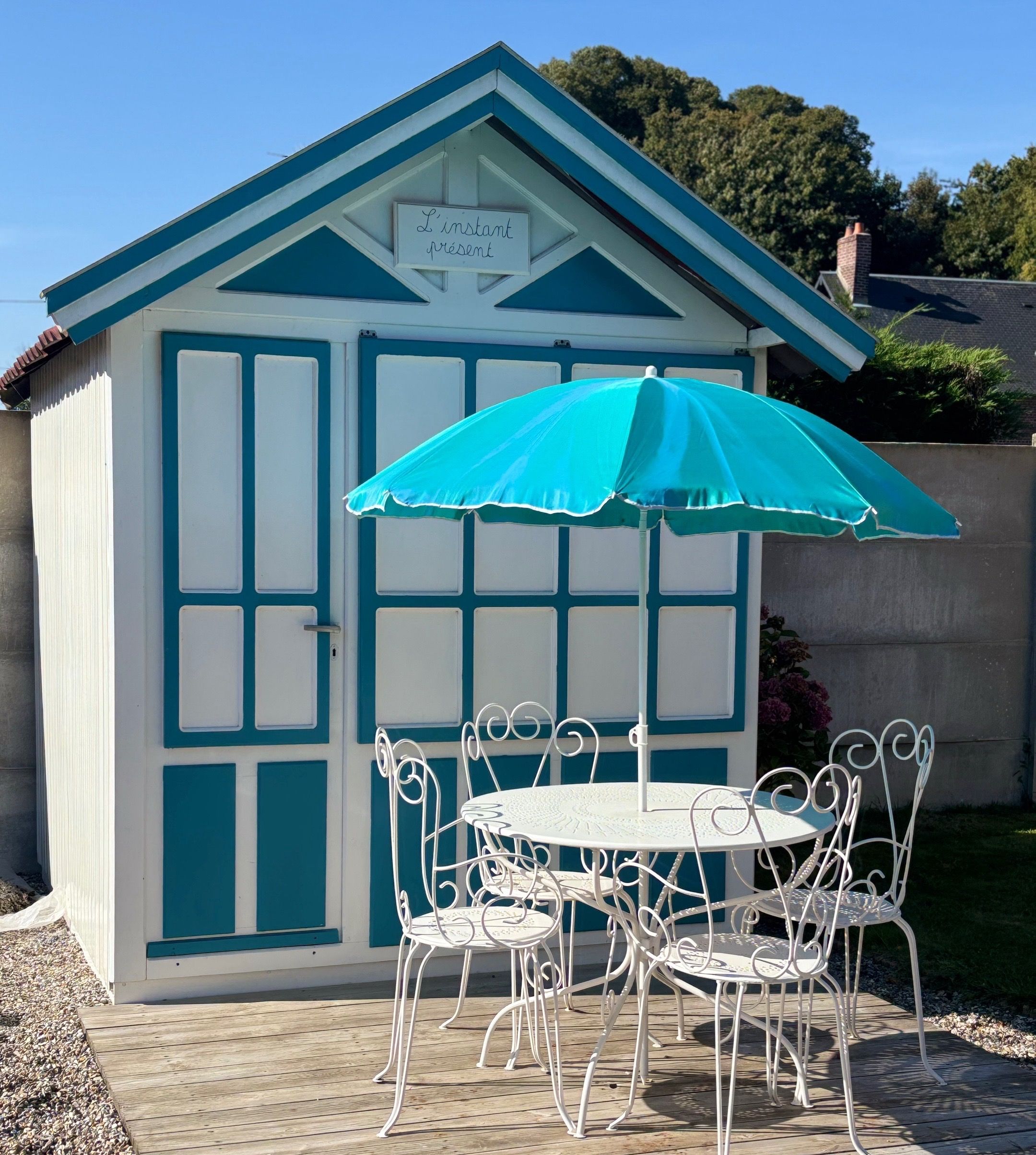 A small, white and blue shed with a sign reading "L'instant présent" above the door. Inside, there is a round table with white metal chairs and a blue umbrella.