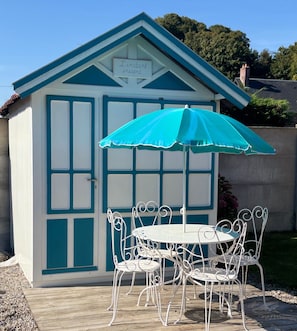 A small, white and blue shed with a sign reading "L'instant présent" above the door. Inside, there is a round table with white metal chairs and a blue umbrella.