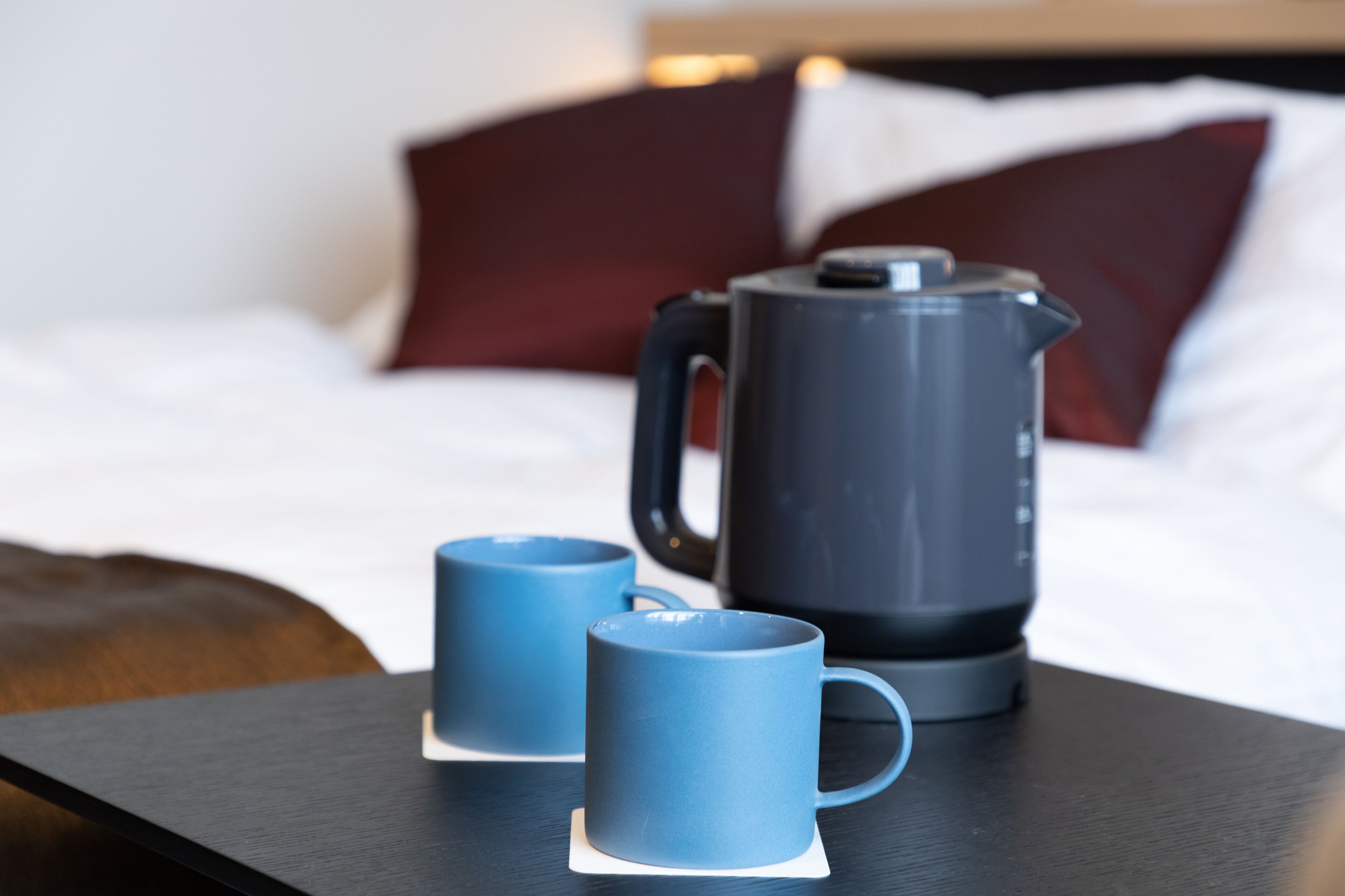 Una cama con sábanas blancas y un cabecero oscuro, una cafetera negra y dos tazas azules sobre una mesa de madera.