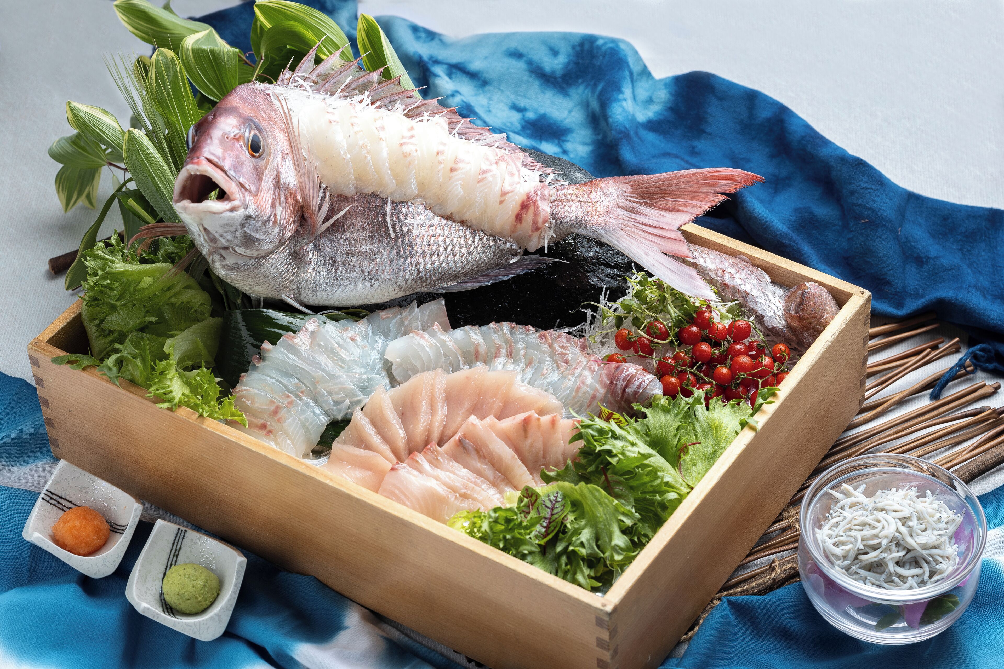 Una caja de madera con diversos productos del mar, como pescado y mariscos, junto con recipientes pequeños de salsas.