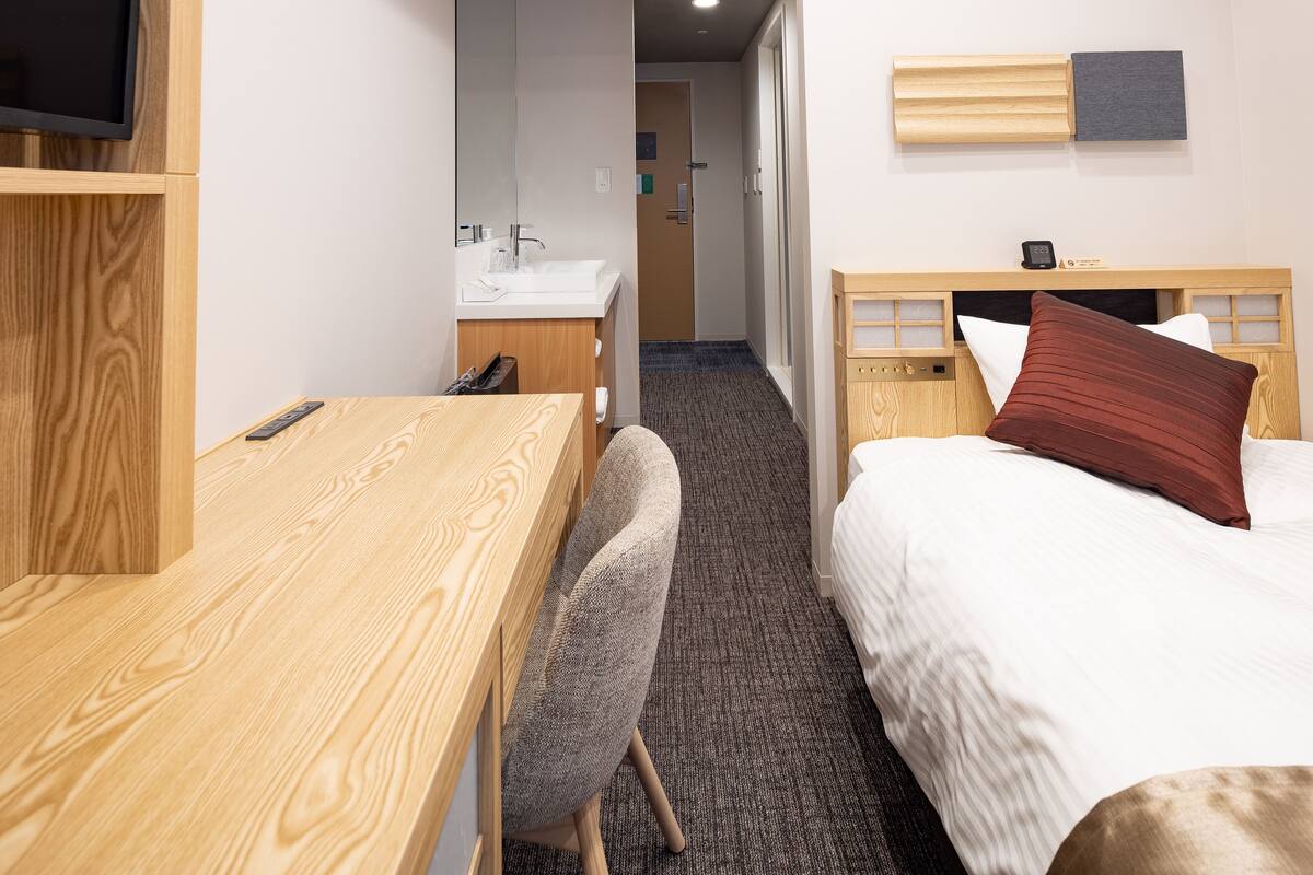 Een hotelkamer met een houten bureau, een stoel, een bed met een rood kussen, een nachtkastje, een spiegel en een televisie aan de muur.