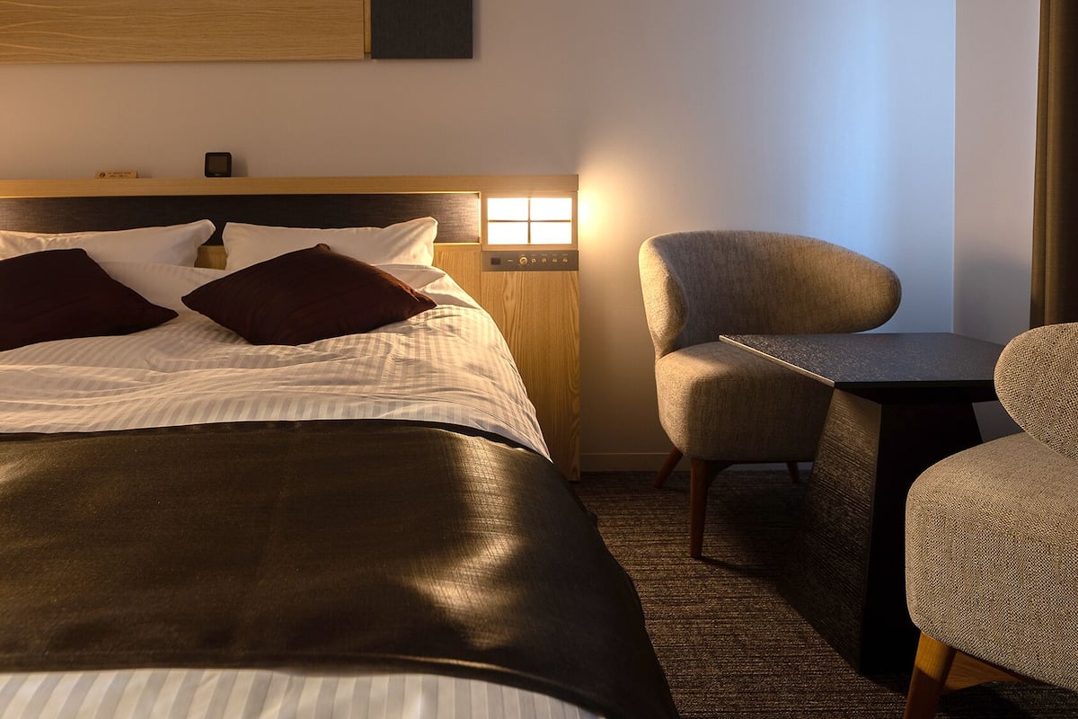 Een hotelkamer met een bed, twee stoelen, een tafeltje en een nachtkastje met een lamp.