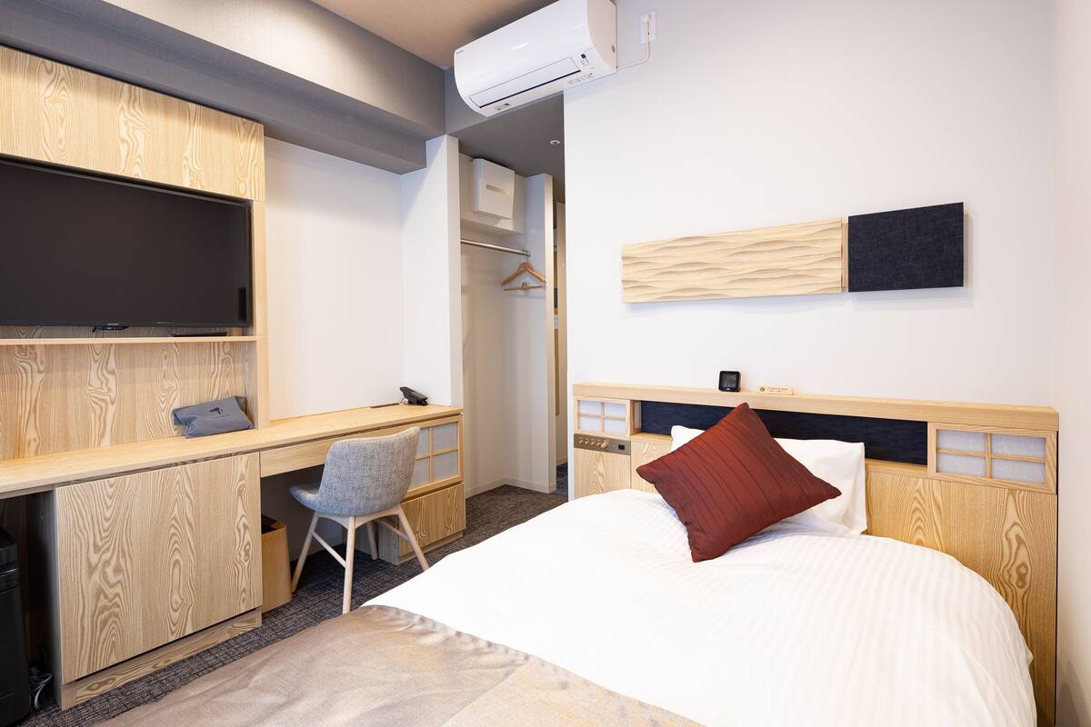 Een hotelkamer met een houten bureau, een stoel, een bed met een rode kussen, een televisie en een garderobe.