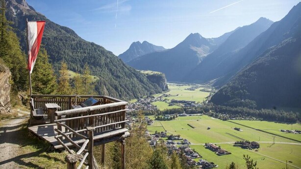 Eine Berglodge mit Holzbalkon und Wimpel, mit Blick auf ein Tal mit Häusern und blauem Himmel.