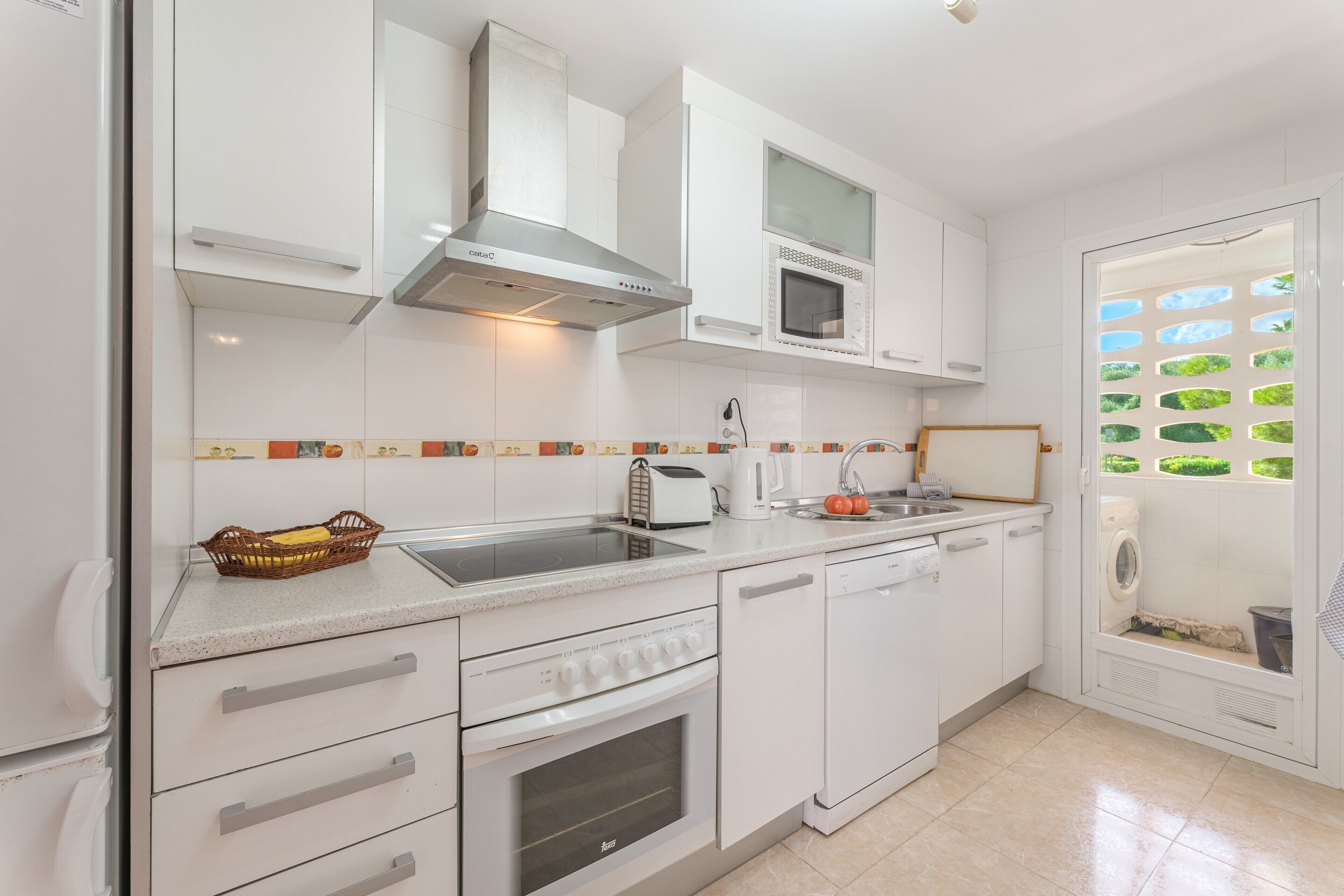 A modern kitchen with white cabinetry, a stainless steel range hood, a built-in microwave, a dishwasher, and a window with a view of greenery.