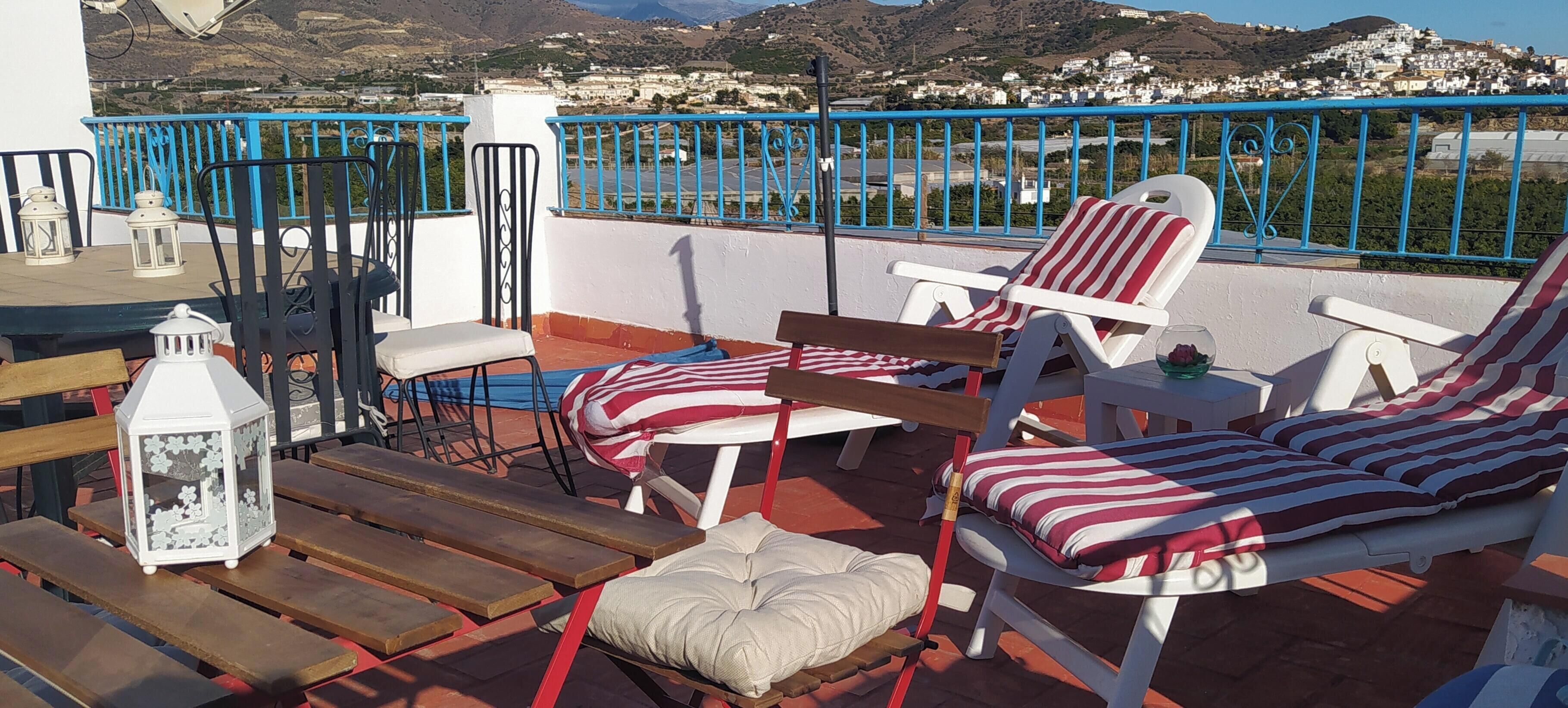 A terrace with outdoor furniture, a view of a coastal town, and a blue railing.