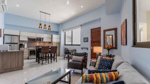 A modern living room with a sofa, armchairs, and a glass coffee table. A kitchen with a dining area is visible in the background. - 5 min to Los Muertos Beach in front of Cuale River located in Isla Cuale! 1BD Co (Puerto Vallarta)