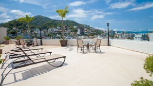 A rooftop terrace with sun loungers, a table, and chairs, overlooking a cityscape with buildings and a hill. - 5 min to Los Muertos Beach in front of Cuale River located in Isla Cuale! 1BD Co (Puerto Vallarta)
