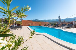 Una piscina con tumbonas y sombrillas, rodeada por una terraza de baldosas y vistas a un paisaje montañoso.