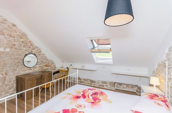 Un dormitorio con techo inclinado, tragaluz, cama con ropa de cama floral, mesita de noche con lámpara y cómoda de madera.