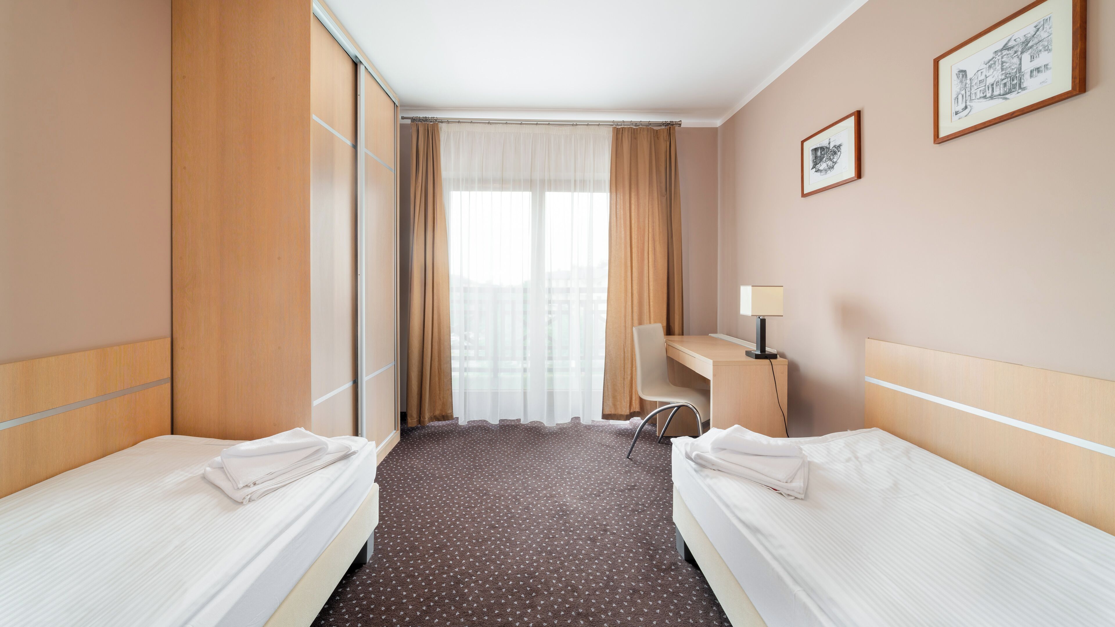 A hotel room with two beds, a desk, a chair, a lamp, and a window with curtains.