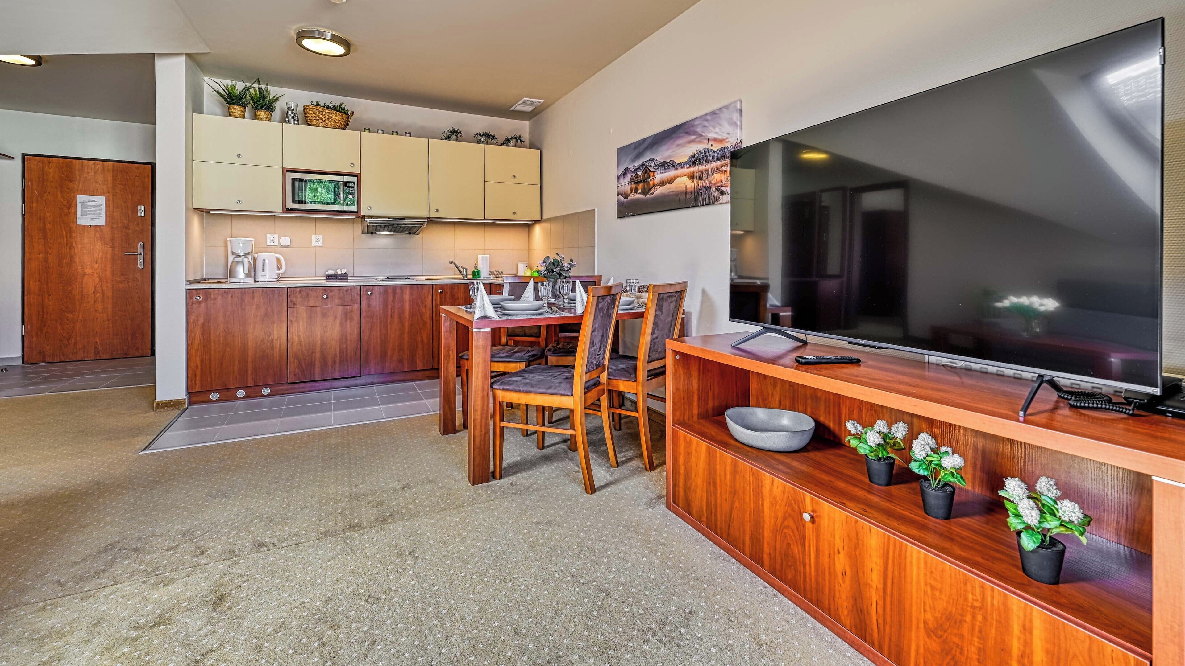 A modern kitchen with wooden cabinets, a dining area with a large TV, and a cozy seating arrangement.