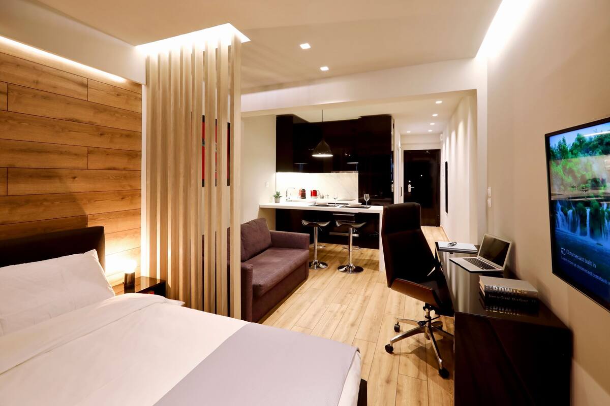 Une chambre d’hôtel avec un lit, un bureau avec un ordinateur portable, une télévision et, au fond, une petite cuisine.