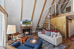A cozy living room with a fireplace, a sofa, and a staircase leading to an upper level. - The Frame by Destination Big Bear : Classic A Frame with Ski Slope Views in Upper Moonridge! (Big Bear Lake)