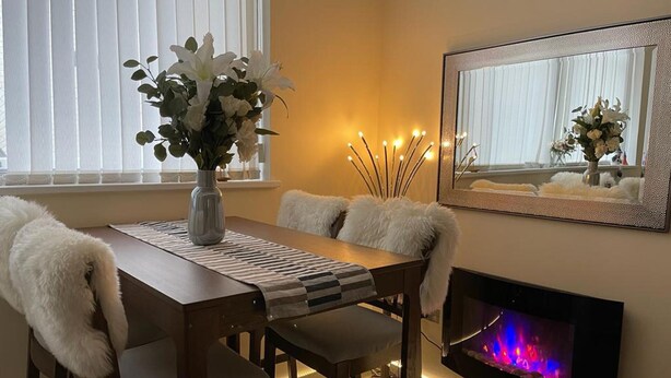 Un coin repas avec une table en bois, des chaises blanches, un vase de fleurs, une cheminée et un miroir au mur.