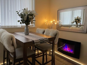 A dining area with a wooden table, white chairs, a vase of flowers, a fireplace, and a mirror on the wall.