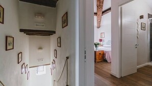 A hallway with framed pictures on the walls, wooden beams on the ceiling, and a door leading to a bedroom with a bed and a dresser.