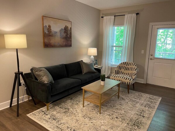 Ein Wohnzimmer mit einem dunkelgrünen Sofa, einem Holztisch, einem gemusterten Teppich, einem weißen Sessel und einem Bild an der Wand.