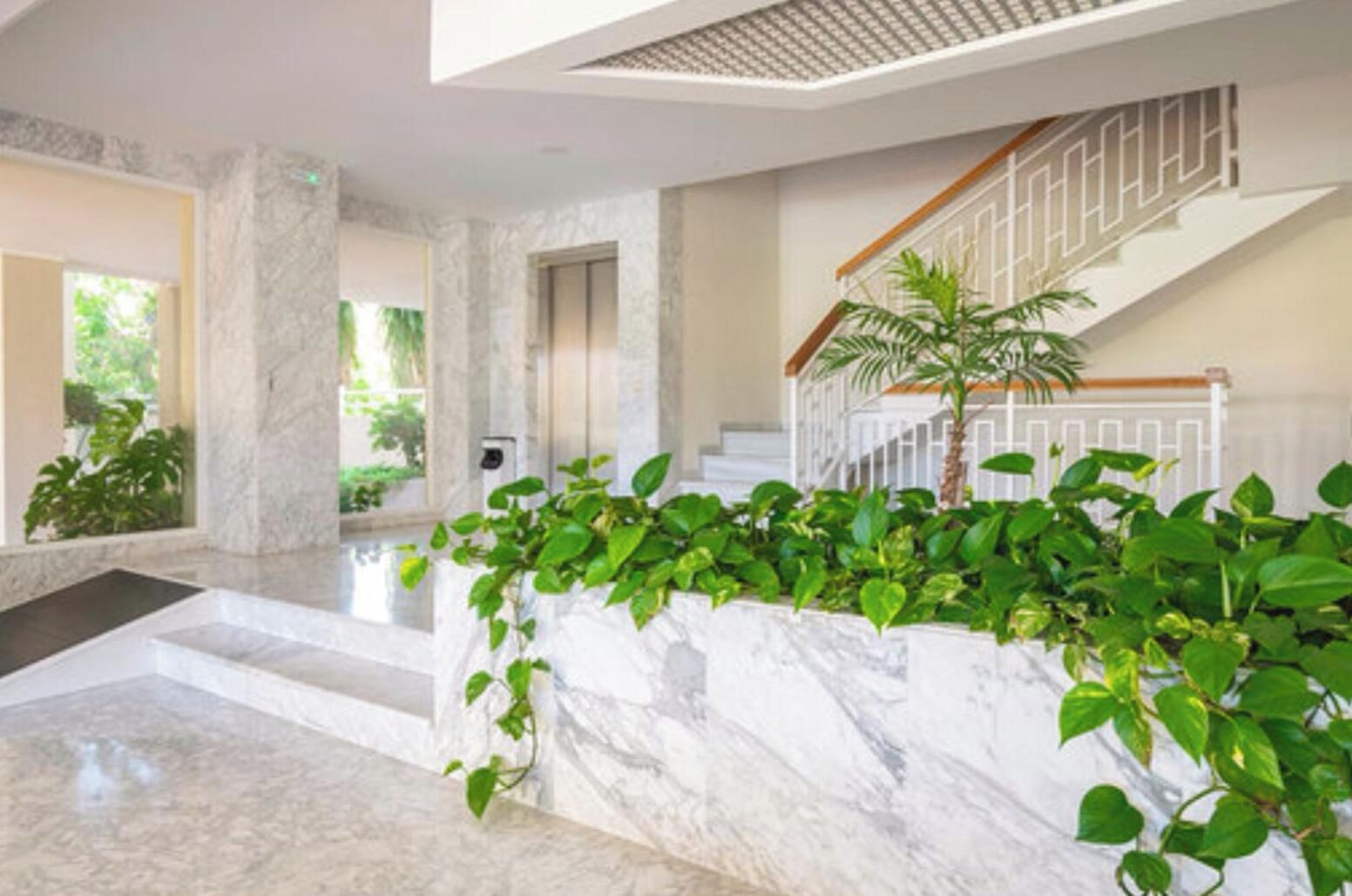 A spacious lobby with a marble floor, a staircase with a wooden handrail, and lush green plants.