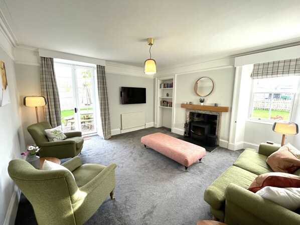 A living room with green armchairs, a pink ottoman, a fireplace, a television, and a bookshelf. - Garden House - Brodie Castle (Forres)