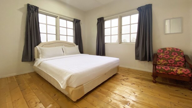 A bedroom with a bed, two windows with curtains, a wooden floor, a ceiling fan, and a chair with a floral pattern. - OYO Home 90329 Bridge House Homestay (Ranau)