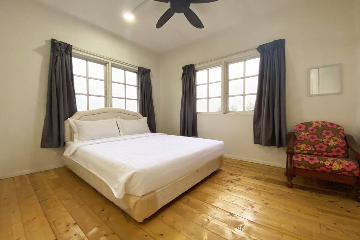 A bedroom with a bed, two windows with curtains, a wooden floor, a ceiling fan, and a chair with a floral pattern.