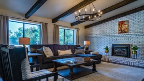 A living room with a dark sofa, a wooden coffee table, and a brick fireplace. - Spa + Fire Pit + Court | Paso Robles Wine Country | Na Hoku Ranch (Atascadero)
