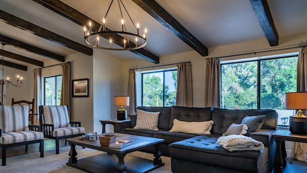 A spacious living room with a dark sofa, armchairs, a coffee table, and a chandelier. Large windows provide natural light and a view of greenery outside. - Spa + Fire Pit + Court | Paso Robles Wine Country | Na Hoku Ranch (Atascadero)