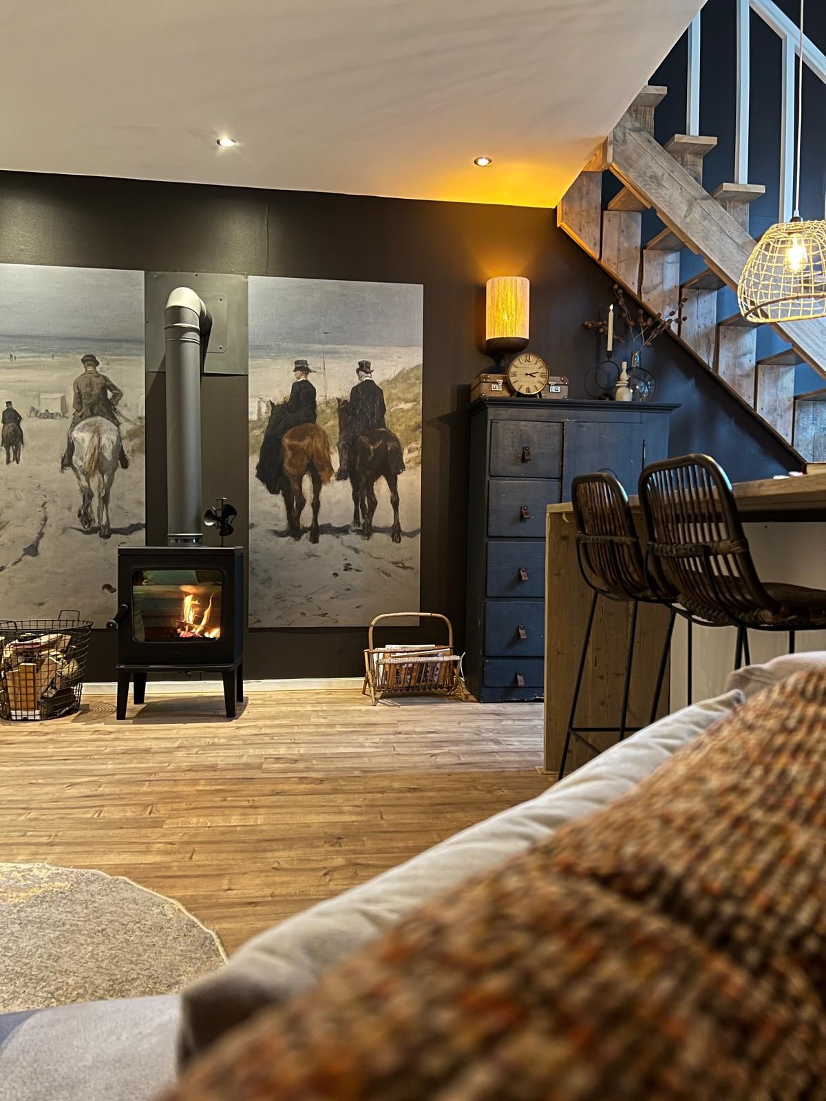 A cozy living room with a wood-burning stove, a large painting of horse riders, and a staircase.