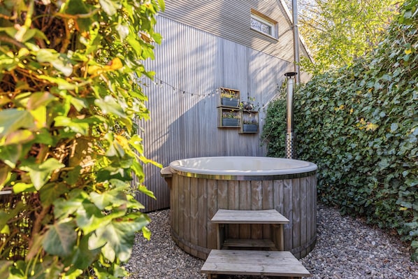 A wooden hot tub with a wooden bench, surrounded by greenery and a wooden fence.